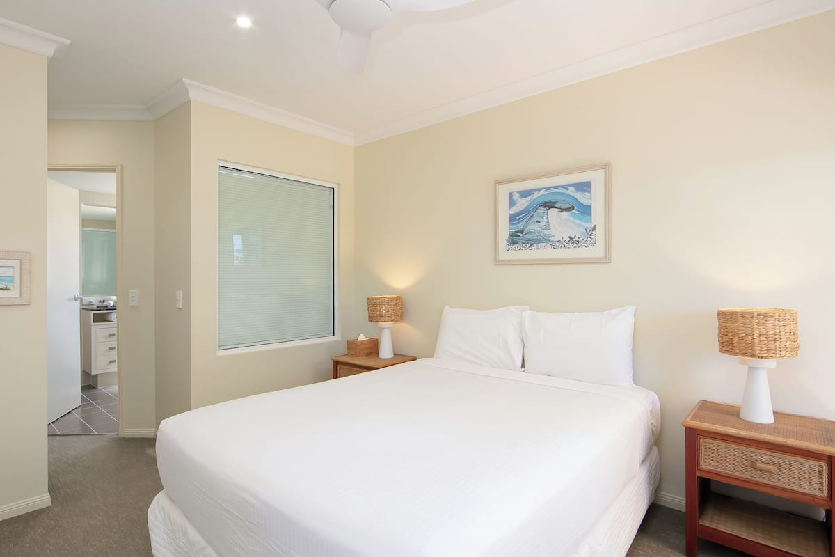 A bedroom with a large bed, a bedside lamp, a picture on the wall, and a window with blinds.