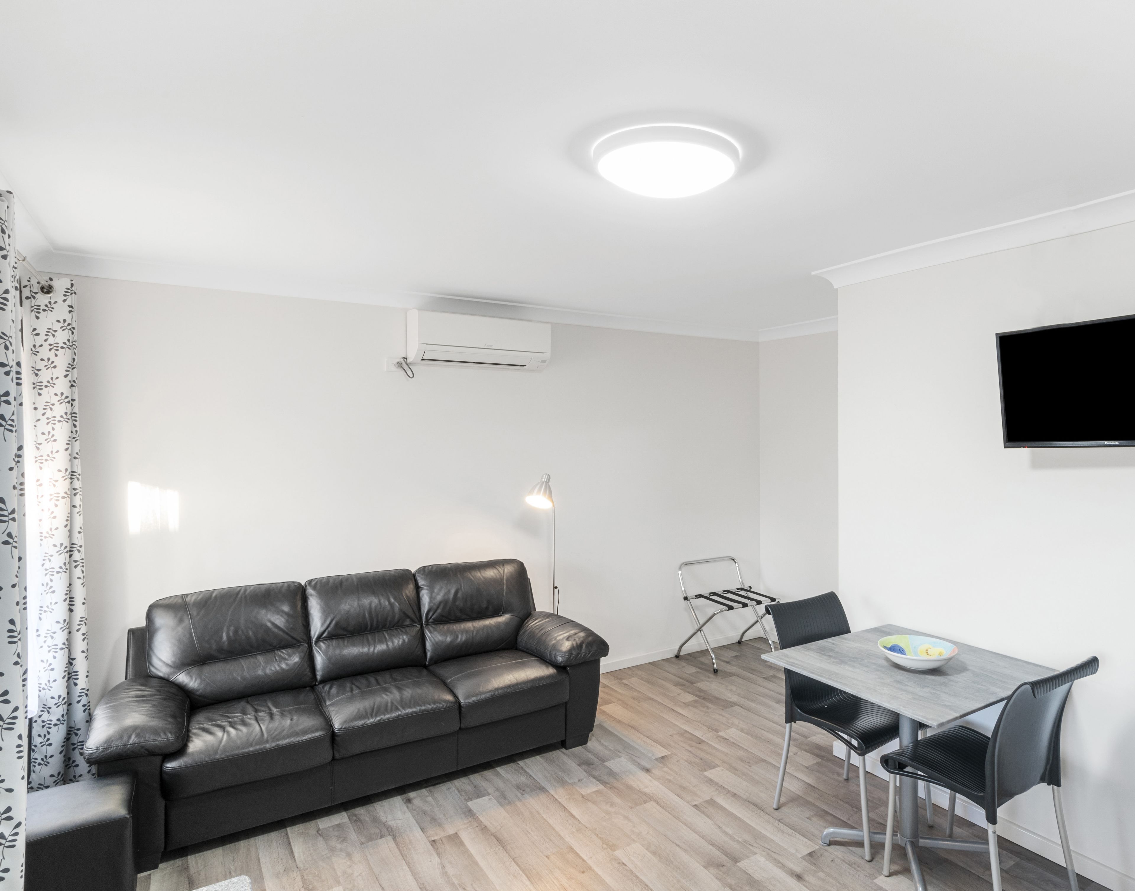 A modern living room with a black leather sofa, a dining table with chairs, a TV mounted on the wall, and an air conditioning unit.