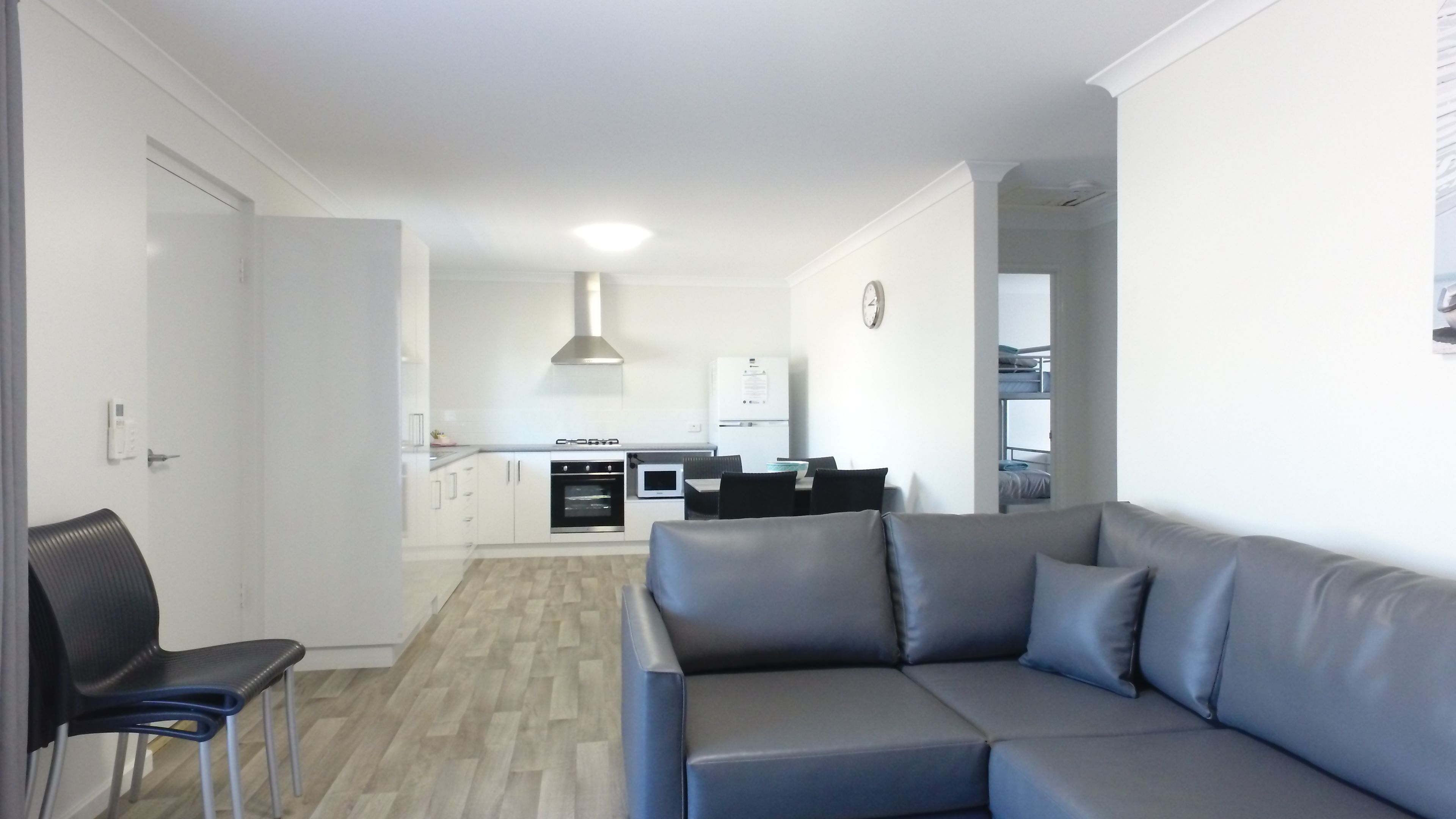 A modern living room with a grey sofa, a dining table with chairs, a kitchen area with white cabinets, and a visible clock on the wall.