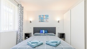 Una cama cuidadosamente arreglada con una colcha a cuadros gris y blanco, dos toallas azules y una almohada azul. Hay dos mesitas de noche con lámparas, una obra de arte en la pared y una ventana con cortina estampada.