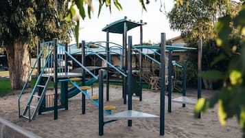 A playground with climbing structures and slides, surrounded by trees and a building in the background.