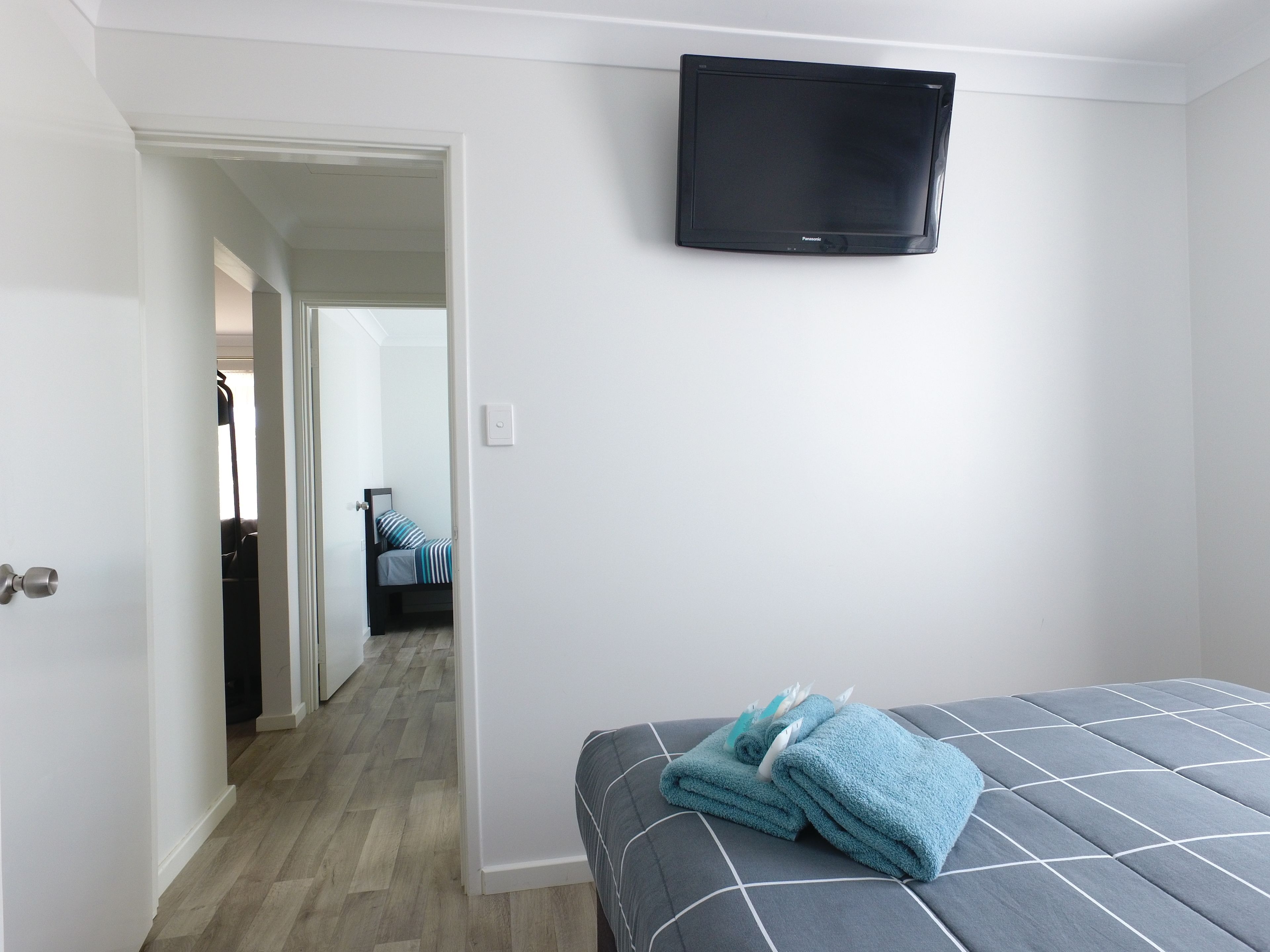 A hotel room with a bed, a flat-screen TV mounted on the wall, and a hallway leading to another room.