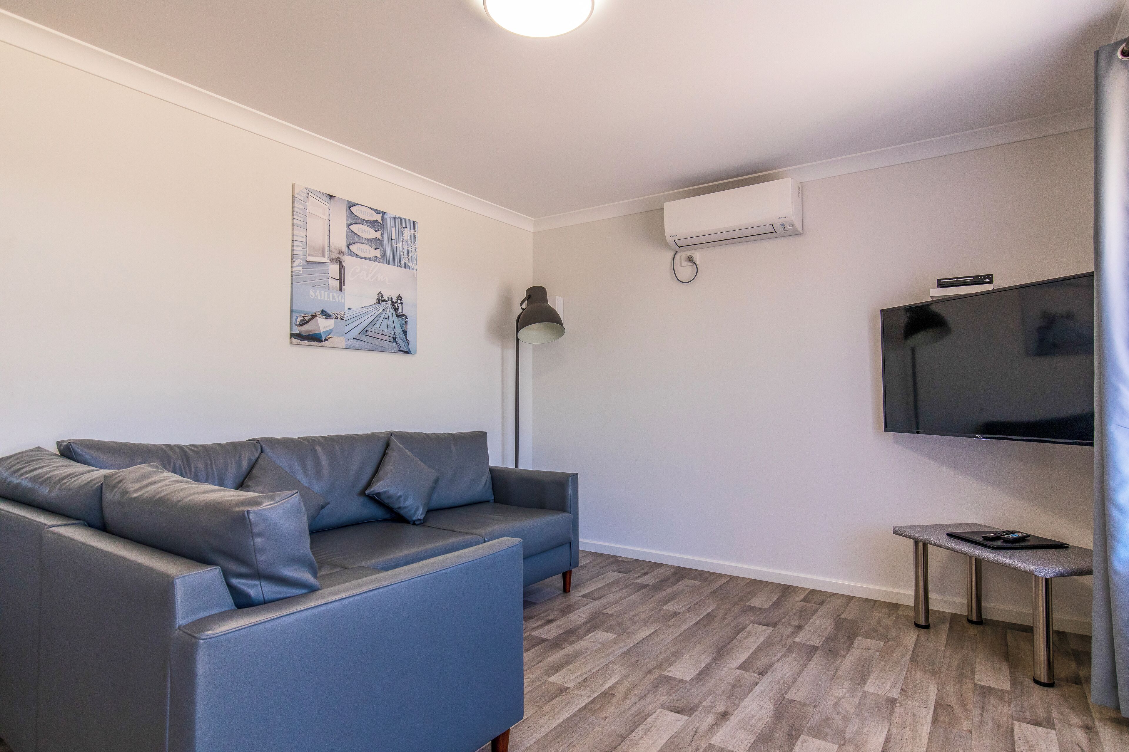 Una sala de estar moderna con un sofá gris, una mesita, una lámpara de pie y un televisor montado.