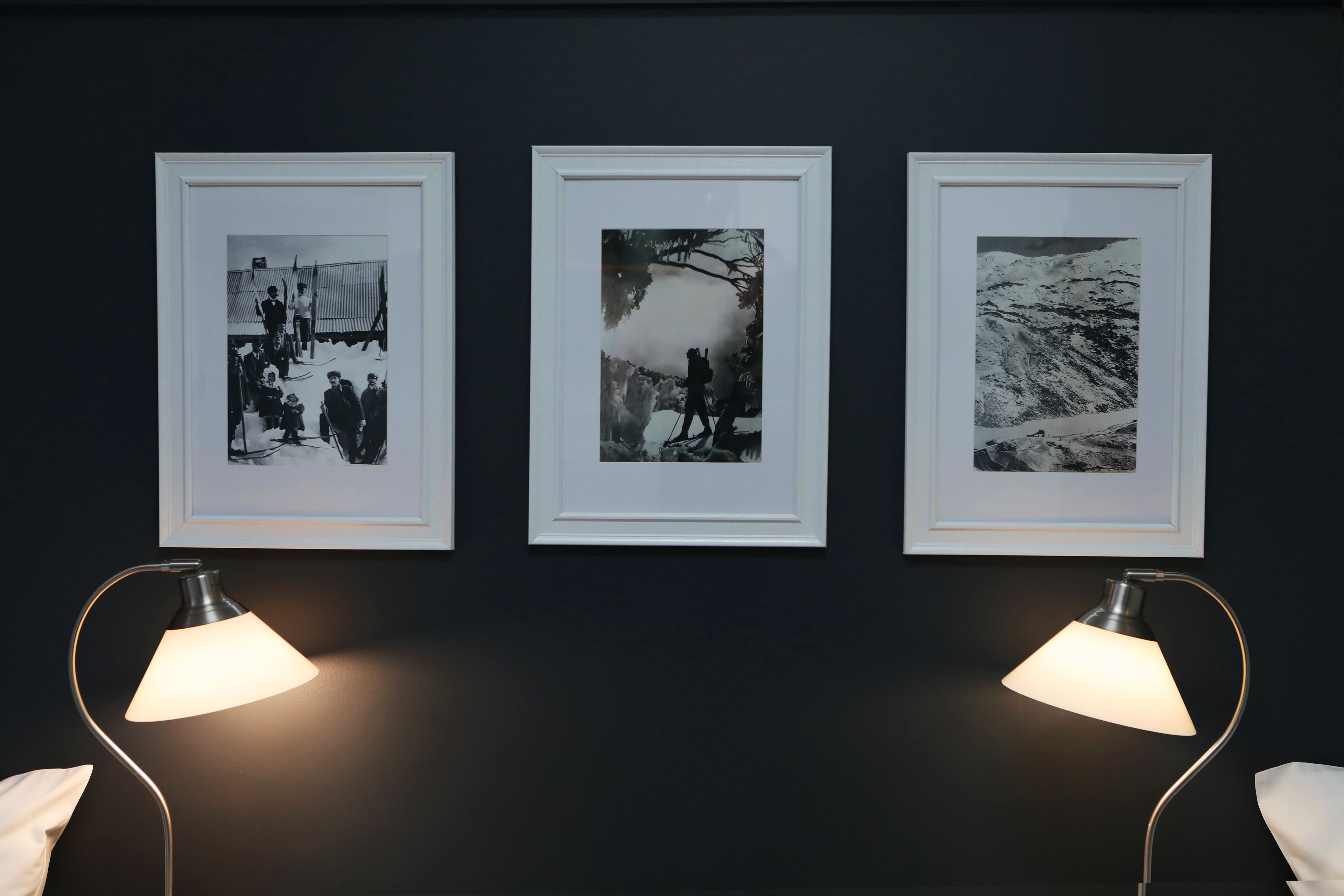 Trois photographies en noir et blanc, encadrées, sont accrochées à un mur sombre, de part et d’autre de deux lampadaires.