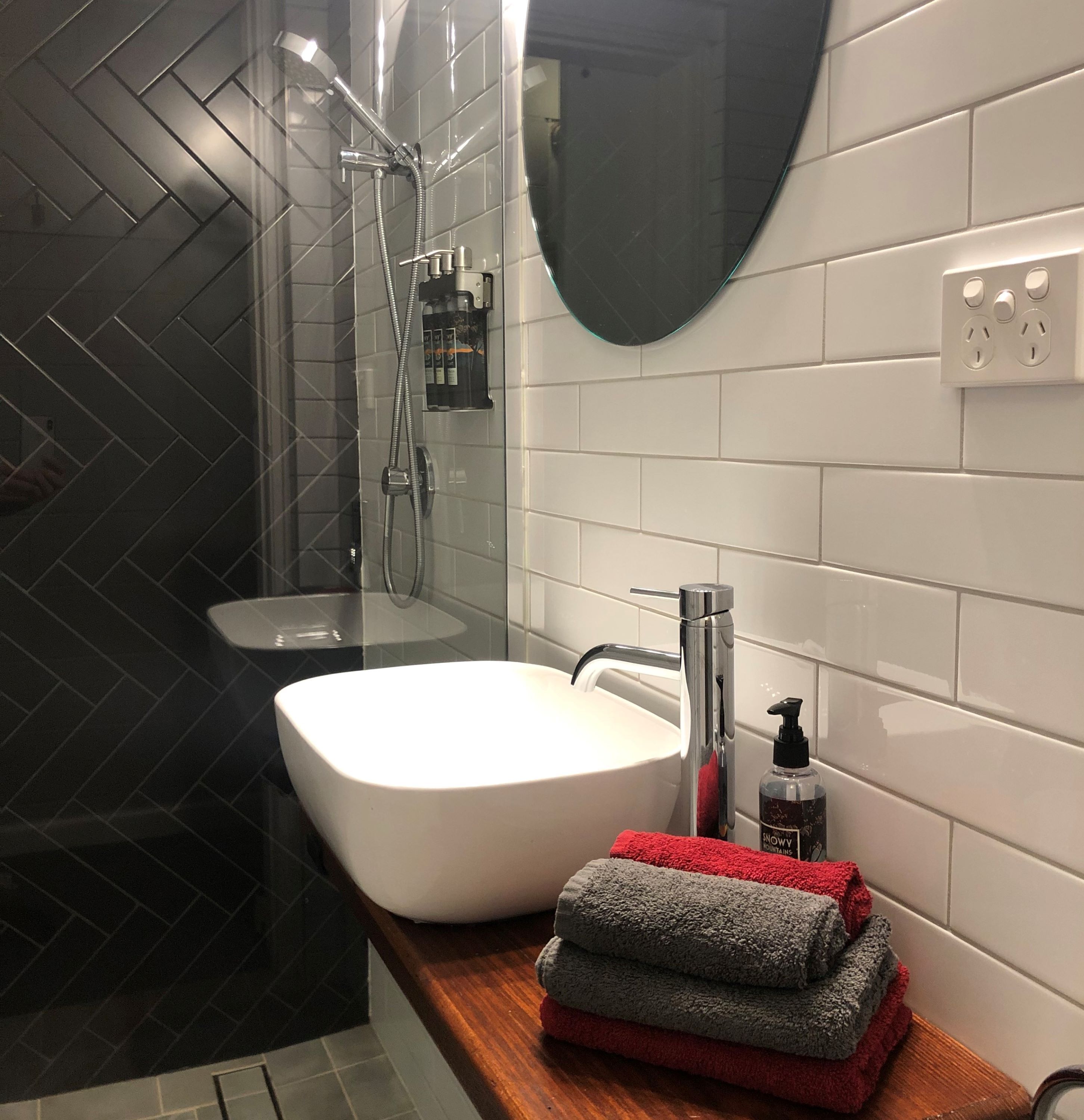 A modern bathroom with a white sink, a shower, and a mirror. There are towels stacked on the counter.