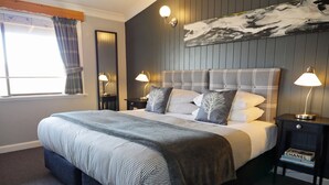 A neatly made bed with a checkered bedspread, two bedside lamps, a nightstand with a book, and a large abstract painting on the wall. - Troldhaugen Lodge - Adults Only (Jindabyne)