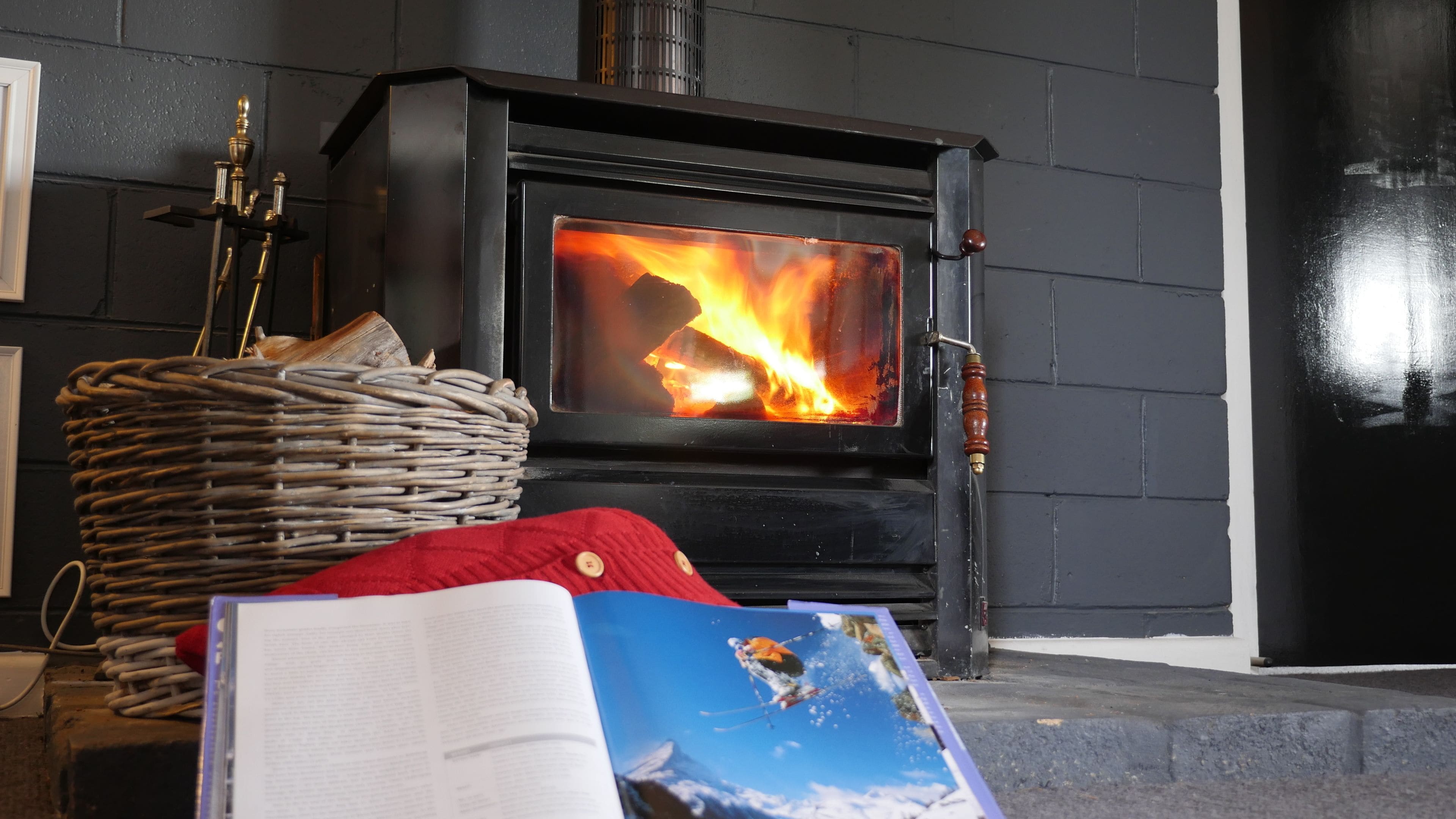 A fireplace with a visible fire, a wicker basket, a red cushion, and a book on a table.