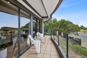 A modern balcony with glass walls, outdoor seating, and a view of the road and greenery. - On The Esplanade Apartments (Torquay)