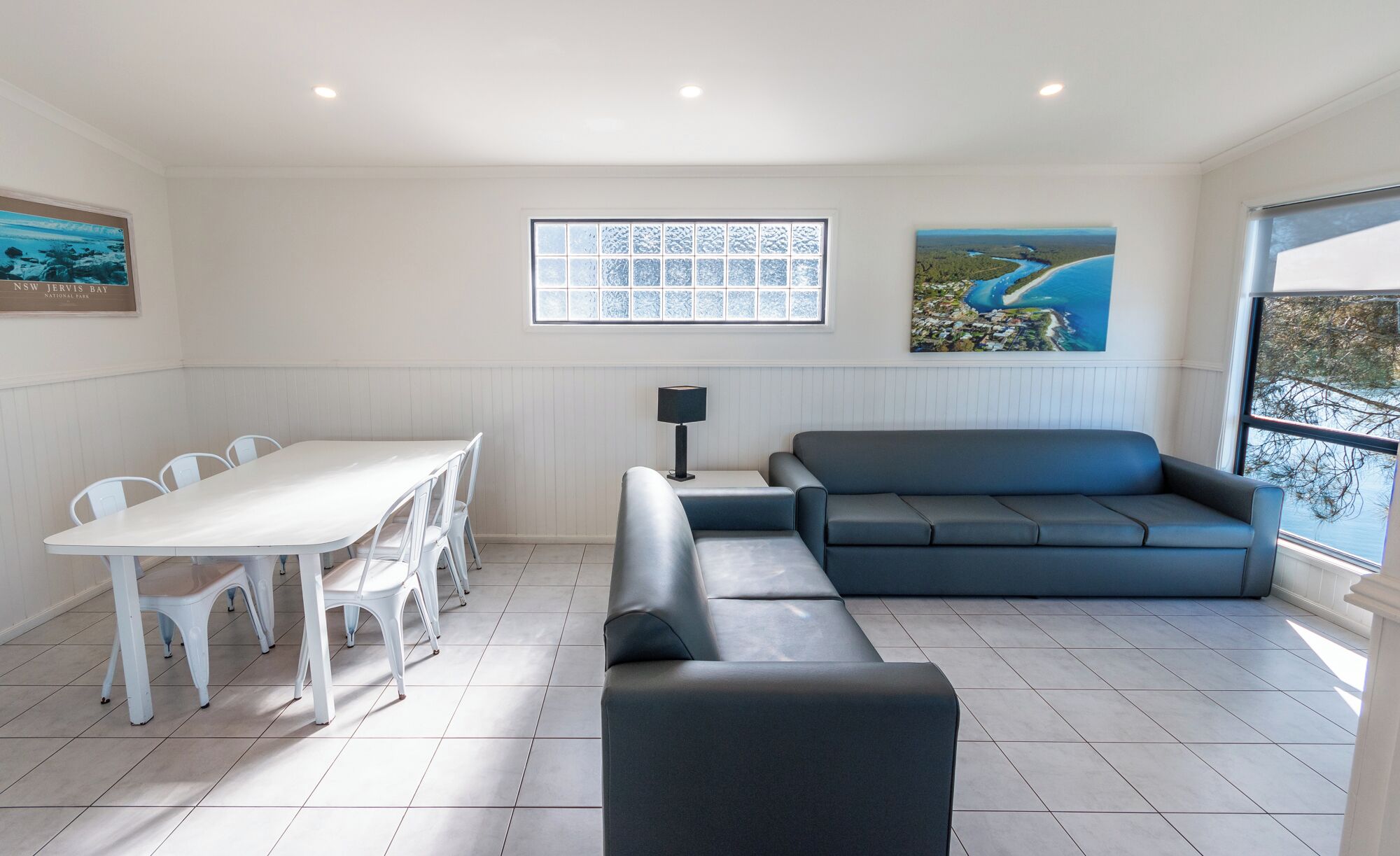 A modern living room with a dining table, chairs, a sofa, and a large window with a view of the sea.