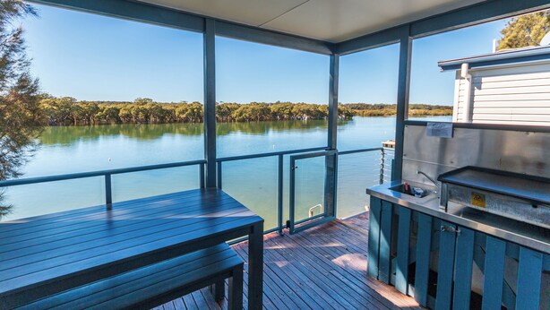 A covered deck with a view of a calm lake and trees, a metal grill, and a small sink.