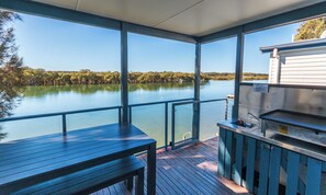 Una terraza techada con vistas a un lago tranquilo y árboles, una parrilla metálica y un lavamanos pequeño.