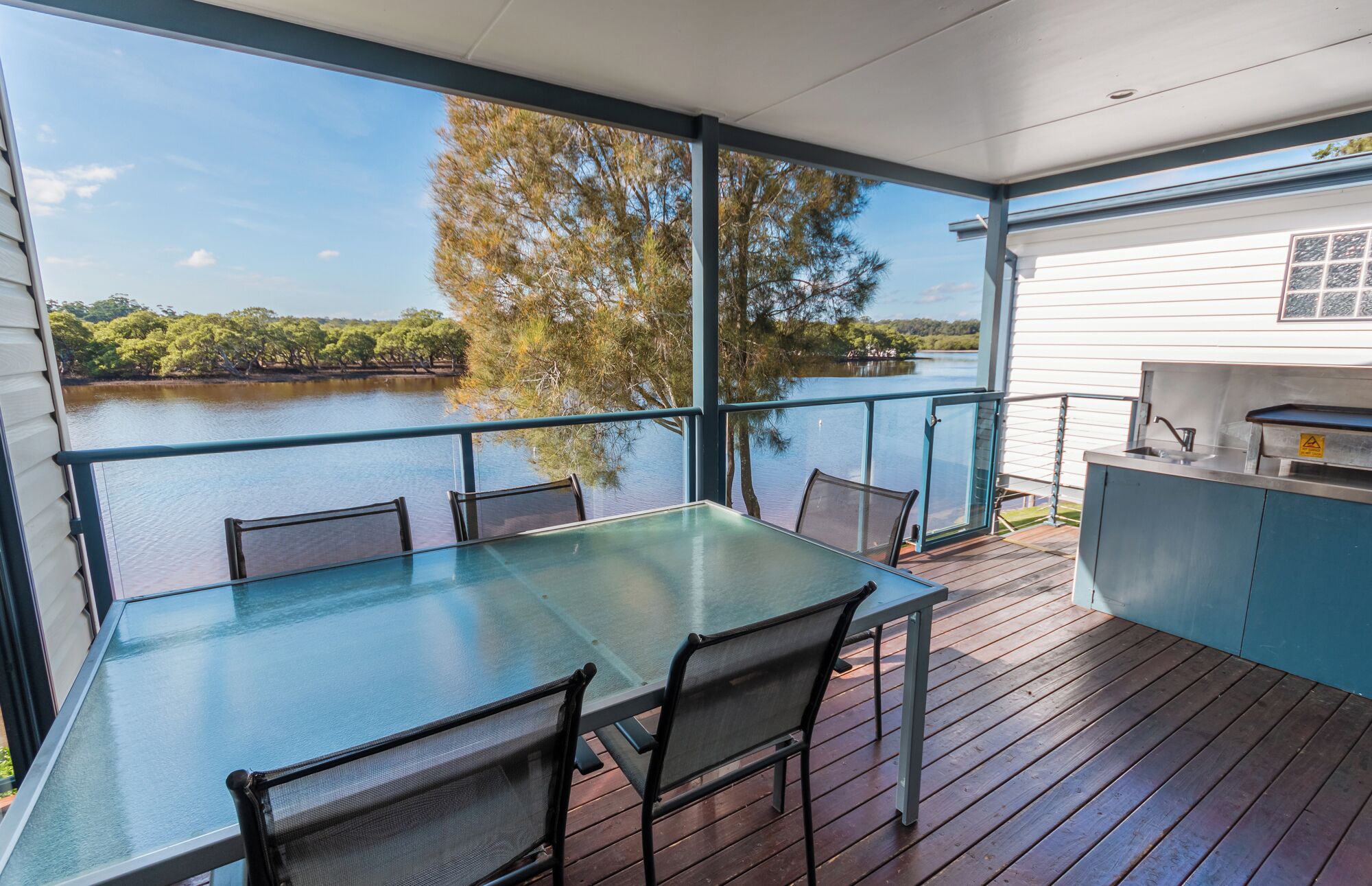 Une terrasse dotée d’une table et de chaises en verre, offrant une vue sur un lac, ainsi qu’un espace barbecue et une vue sur les arbres et l’eau.