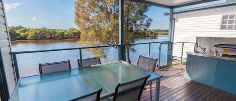 Terraza con mesa y sillas de vidrio con vistas a un lago, zona de barbacoa y vistas a árboles y agua.