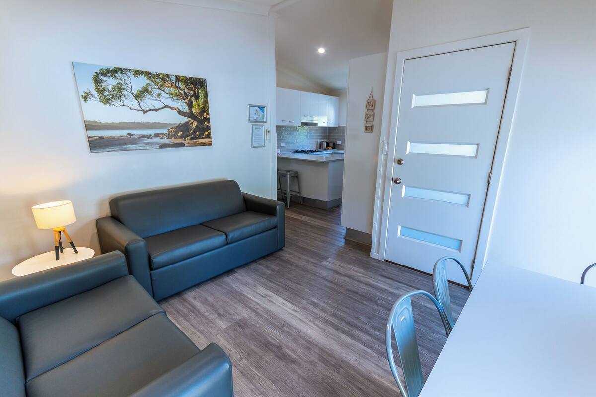 En stue med sofa, et lille bord med lampe, et spiseområde med stol og et køkken i baggrunden.