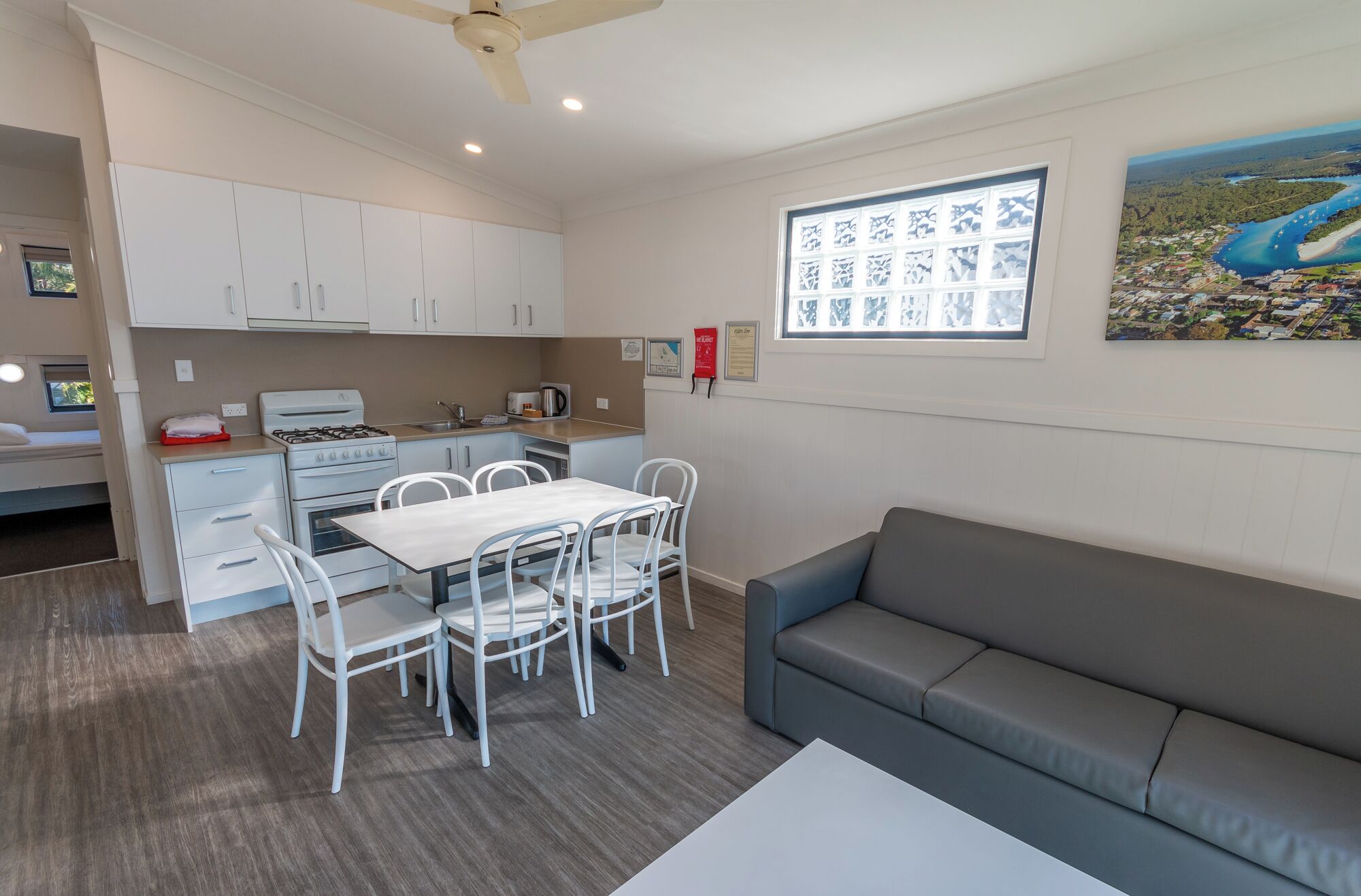 A compact kitchen with white cabinets, a dining table with chairs, a gray sofa, and a painting of a landscape on the wall.