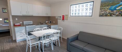 A compact kitchen with white cabinets, a dining table with chairs, a gray sofa, and a painting of a landscape on the wall.