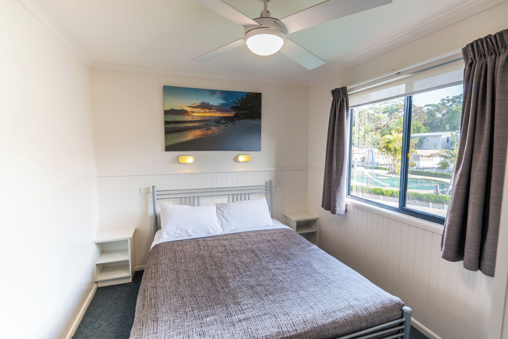 Un dormitorio con cama, ventilador de techo, un cuadro, ventana con cortinas y vistas a una piscina y vegetación.