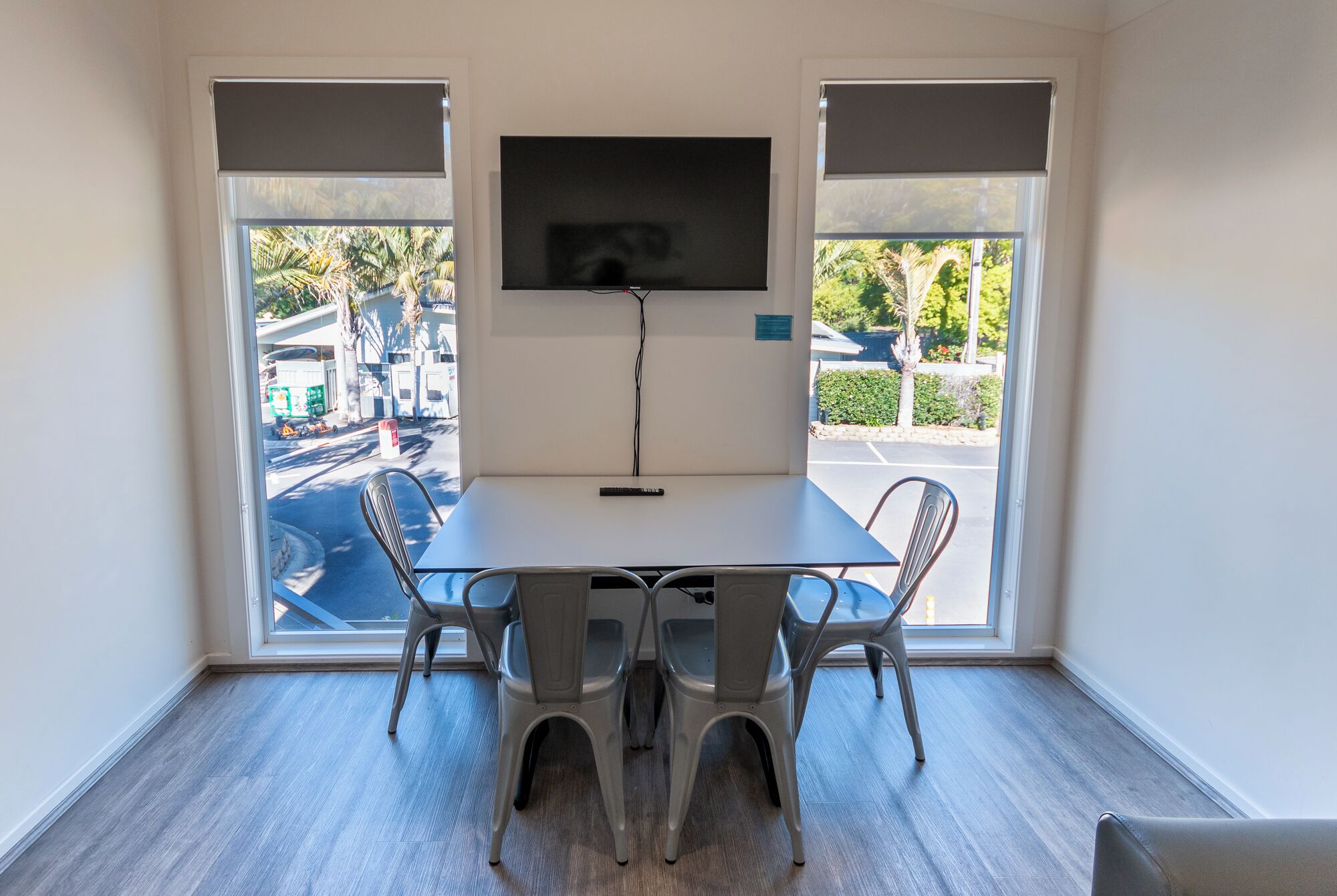 A room with a TV mounted on the wall, a table with four chairs, and large windows with blinds.