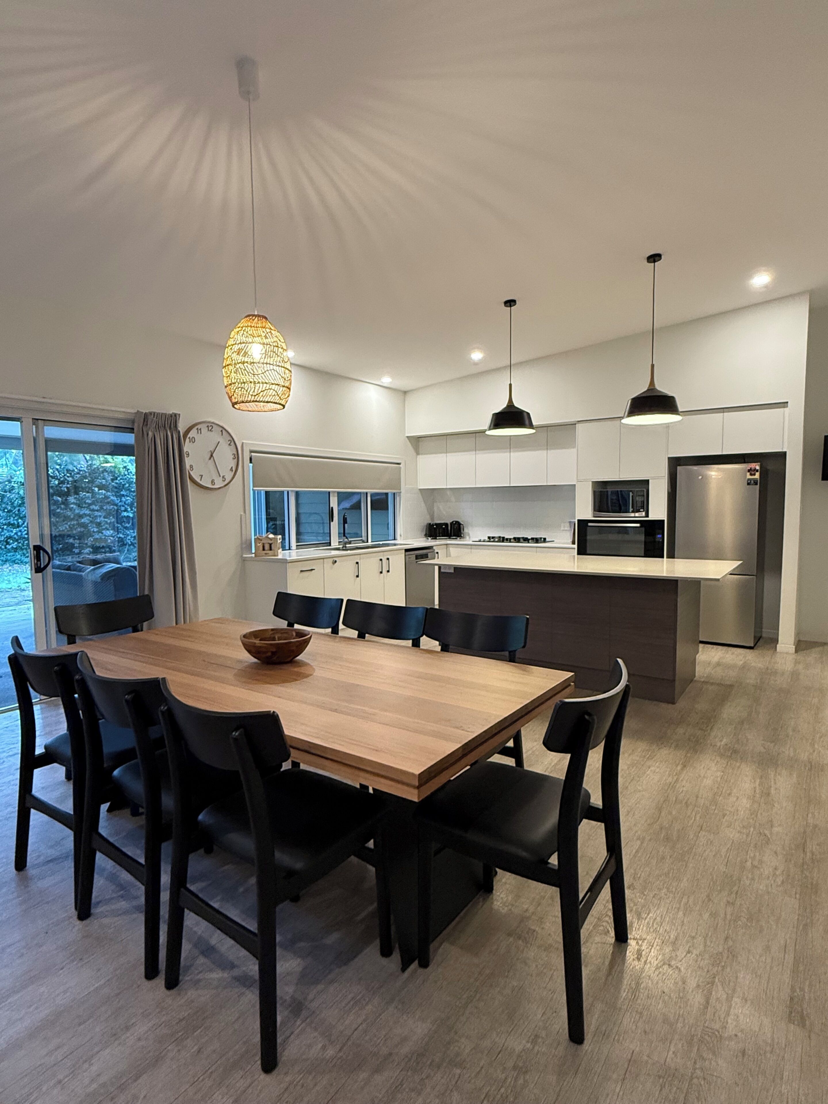 A modern kitchen with a dining area, featuring a wooden table, black chairs, and pendant lighting.