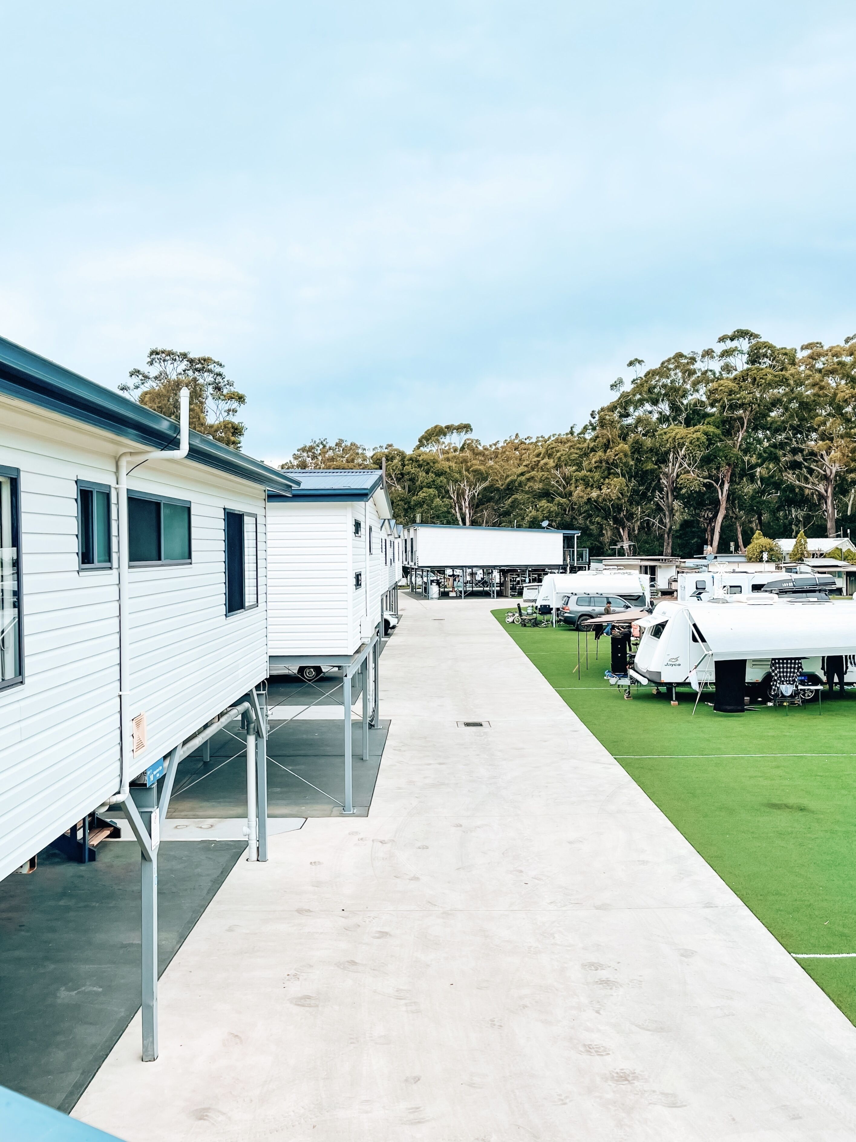 A row of mobile homes with a concrete pathway leading to a grassy area with RVs parked.