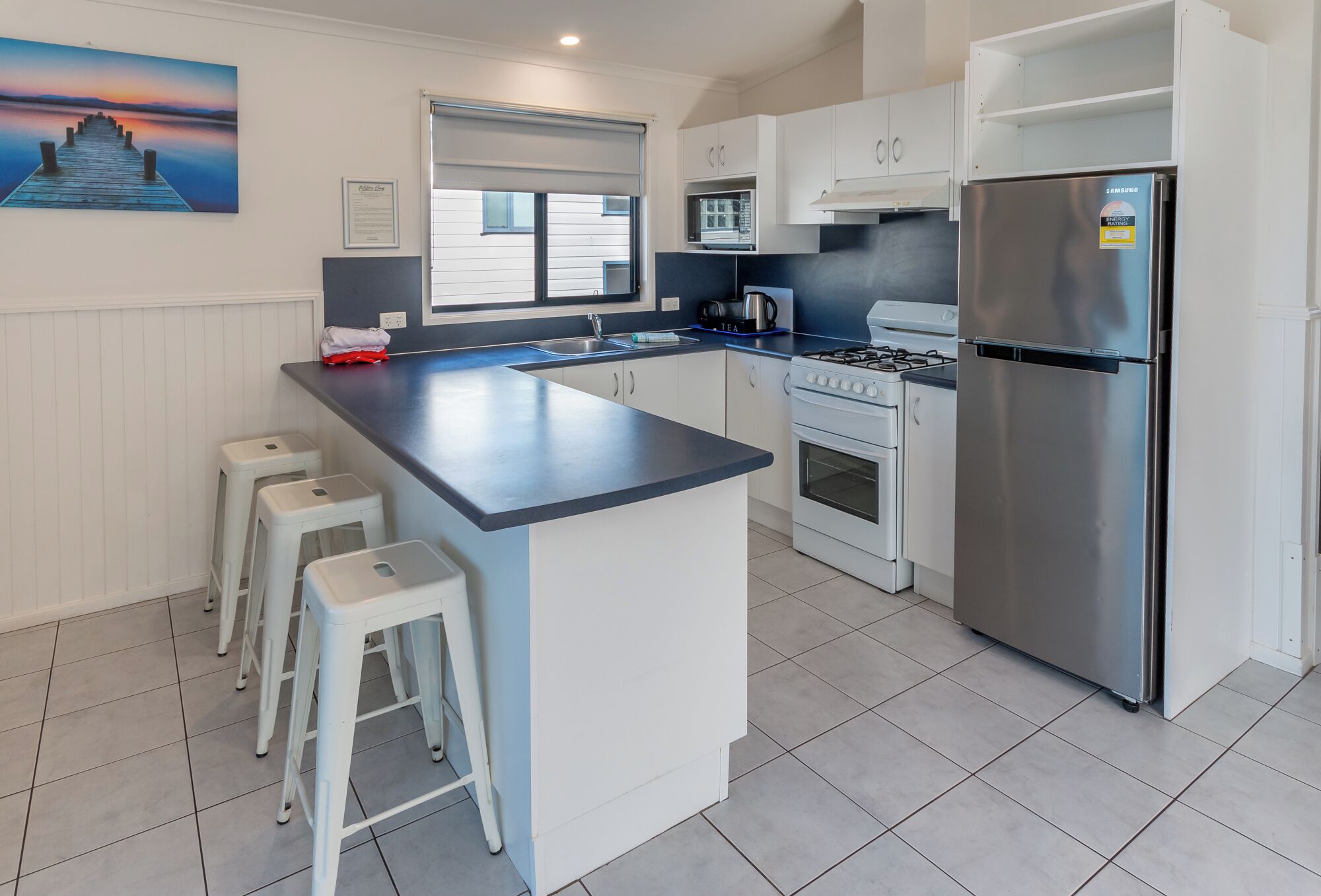 A modern kitchen with a central island, white cabinetry, a stainless steel refrigerator, a gas stove, and a microwave. There are three white stools at the island.