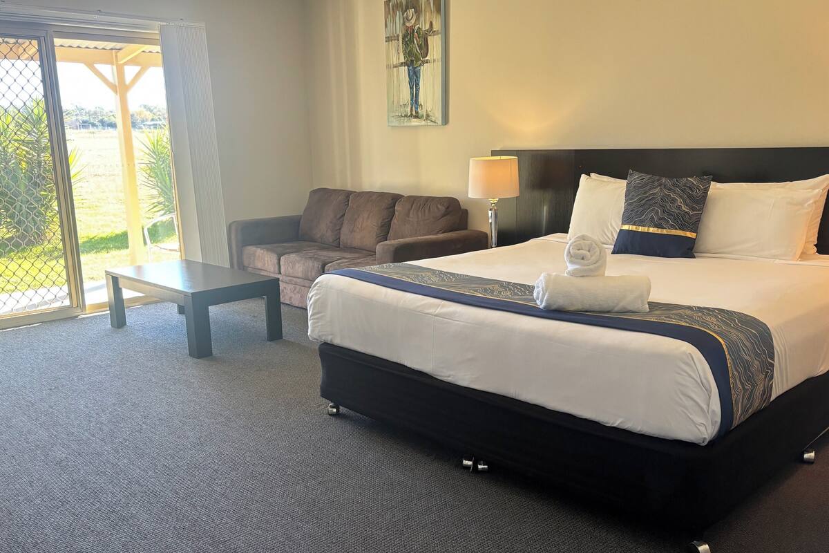A hotel room with a large bed, a sofa, a small table, and a view of the outdoors through a glass door.