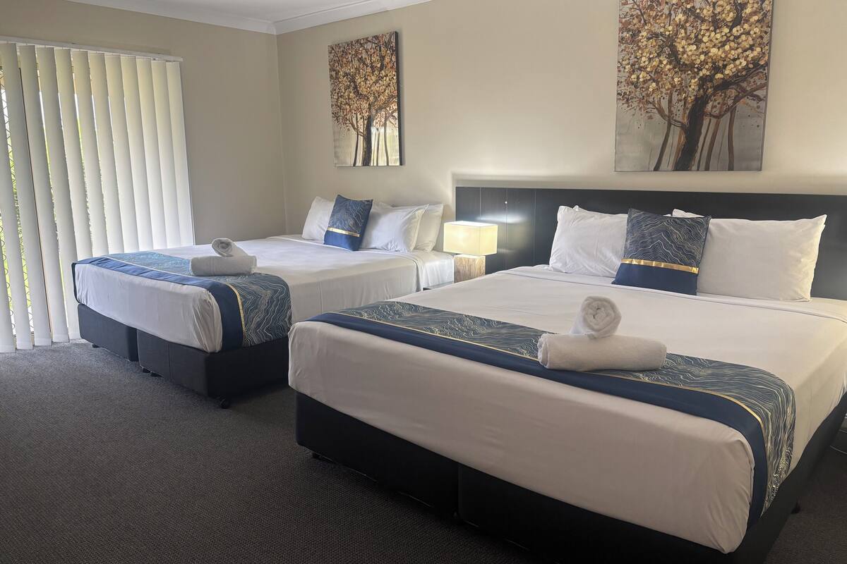 A hotel room with two beds, a headboard, a nightstand, a lamp, and artwork on the walls.