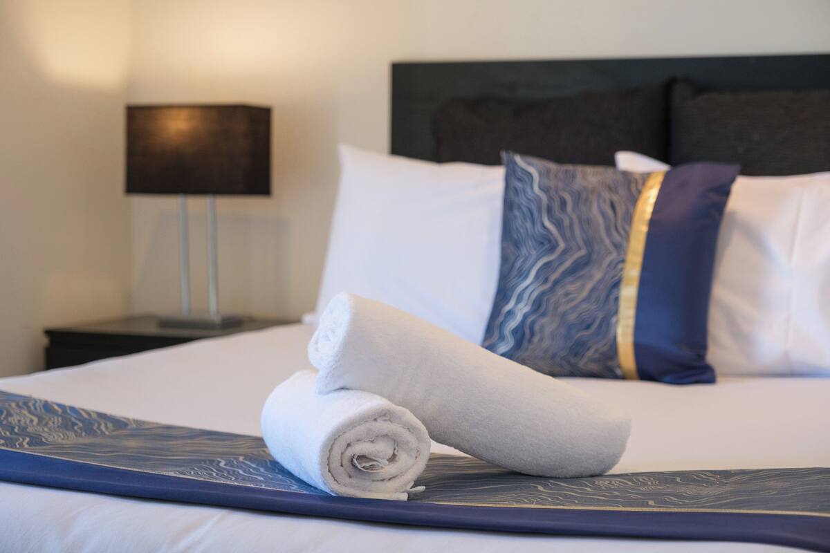 A neatly made bed with a rolled towel, a patterned pillow, and a bedside lamp.