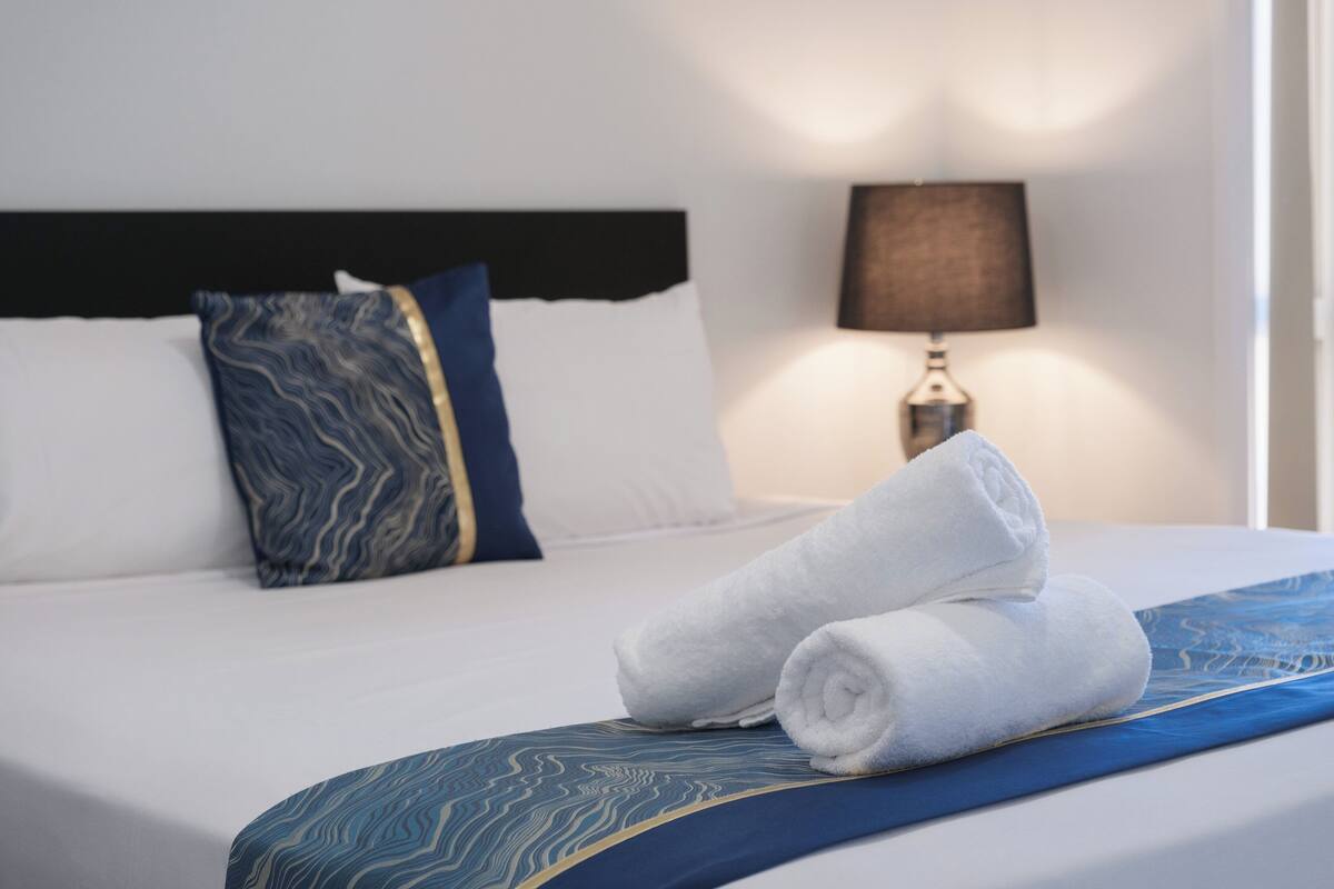 A neatly made bed with white linens, a blue patterned pillow, and a rolled white towel on the bed.