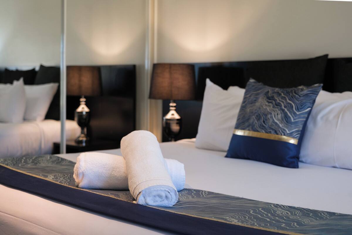 A neatly made bed with rolled towels, a dark headboard, and two bedside lamps.