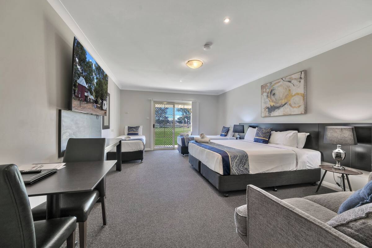 A modern hotel room with a large bed, a desk, a chair, and a sofa. There is a painting on the wall and a window with a view of the outdoors.