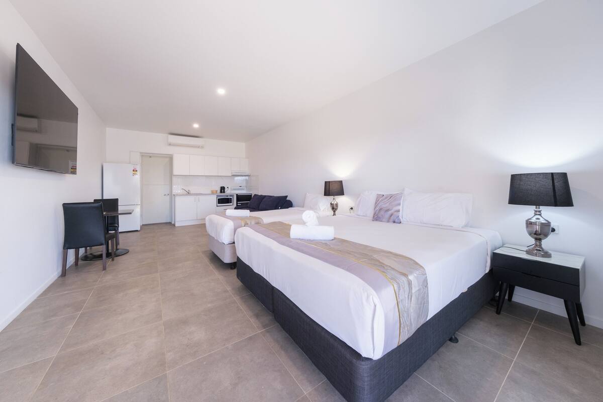 A modern hotel room with a large bed, a desk, a chair, a small kitchenette, and a separate bathroom area.