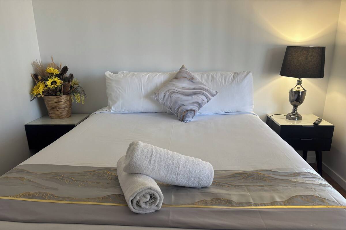 A neatly made bed with white linens, a rolled towel, and a decorative pillow. There is a nightstand with a lamp and a small potted plant beside the bed.