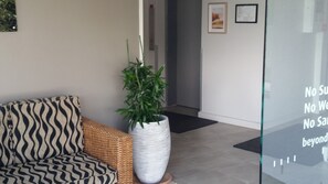 A modern interior with a wicker sofa, a large potted plant, and a glass wall with motivational text. - Amira Apartments (Main Beach)