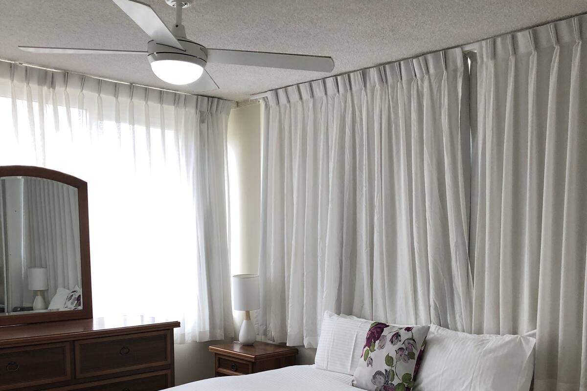 Ein ordentlich bezogenes Bett mit Blumenmuster, ein Nachttisch aus Holz mit Lampe, ein Deckenventilator und leichte, weiße Vorhänge.