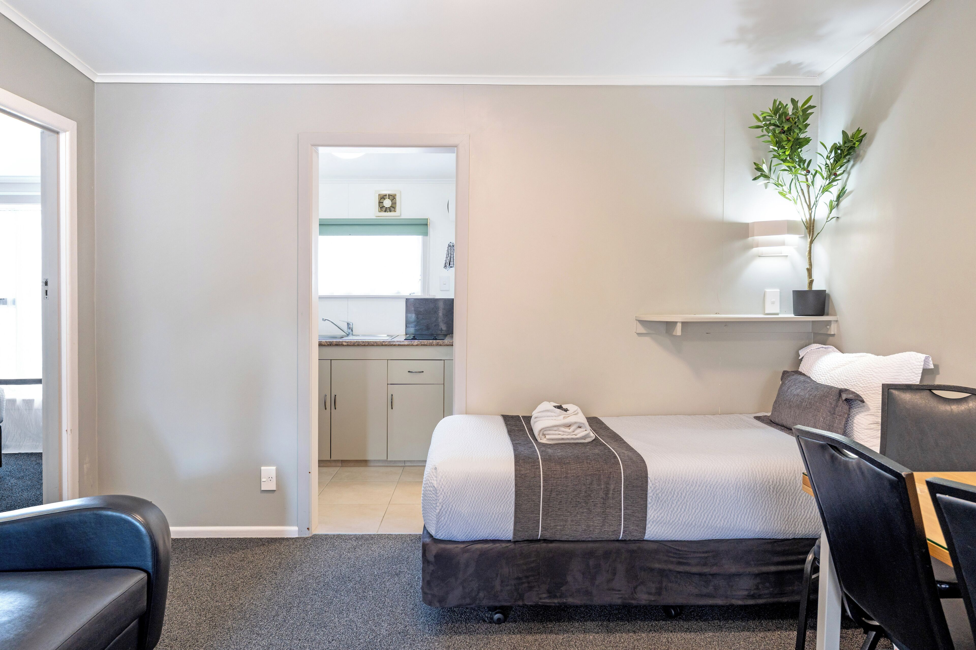 A bedroom with a bed, a desk, a chair, a potted plant, and a view into the kitchen.