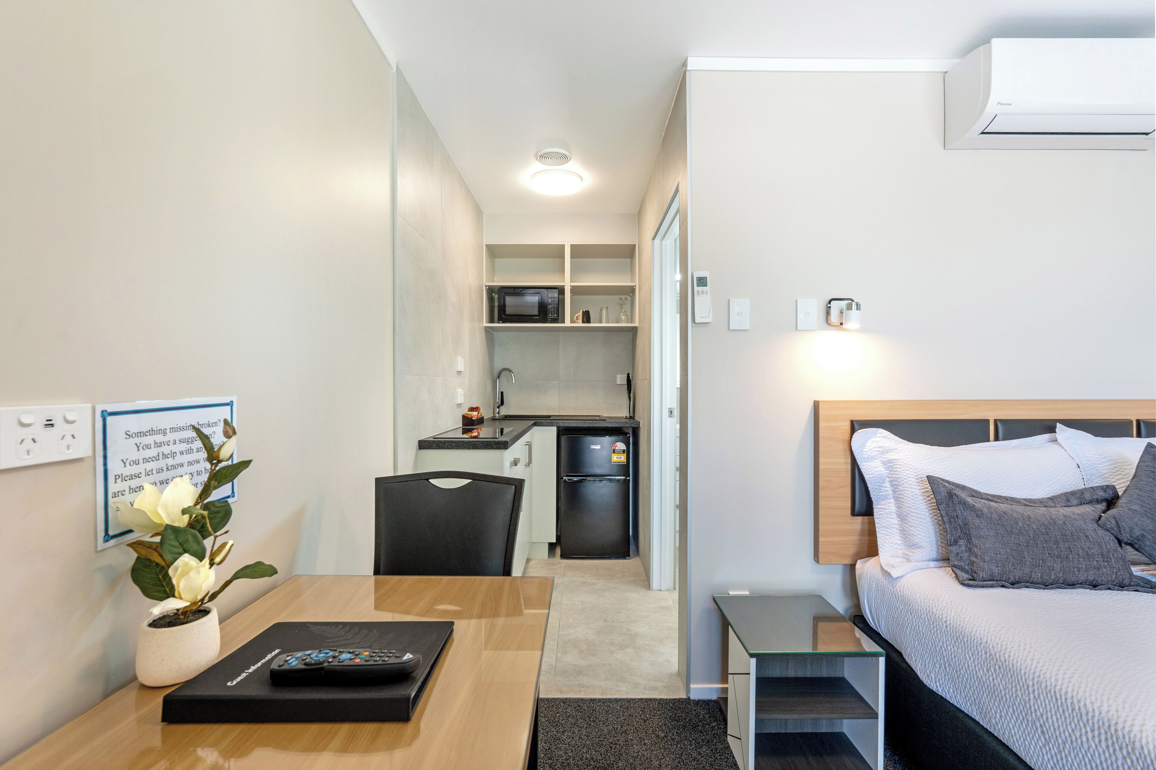 A compact hotel room with a bed, bedside table, a small kitchenette, and a dining area.