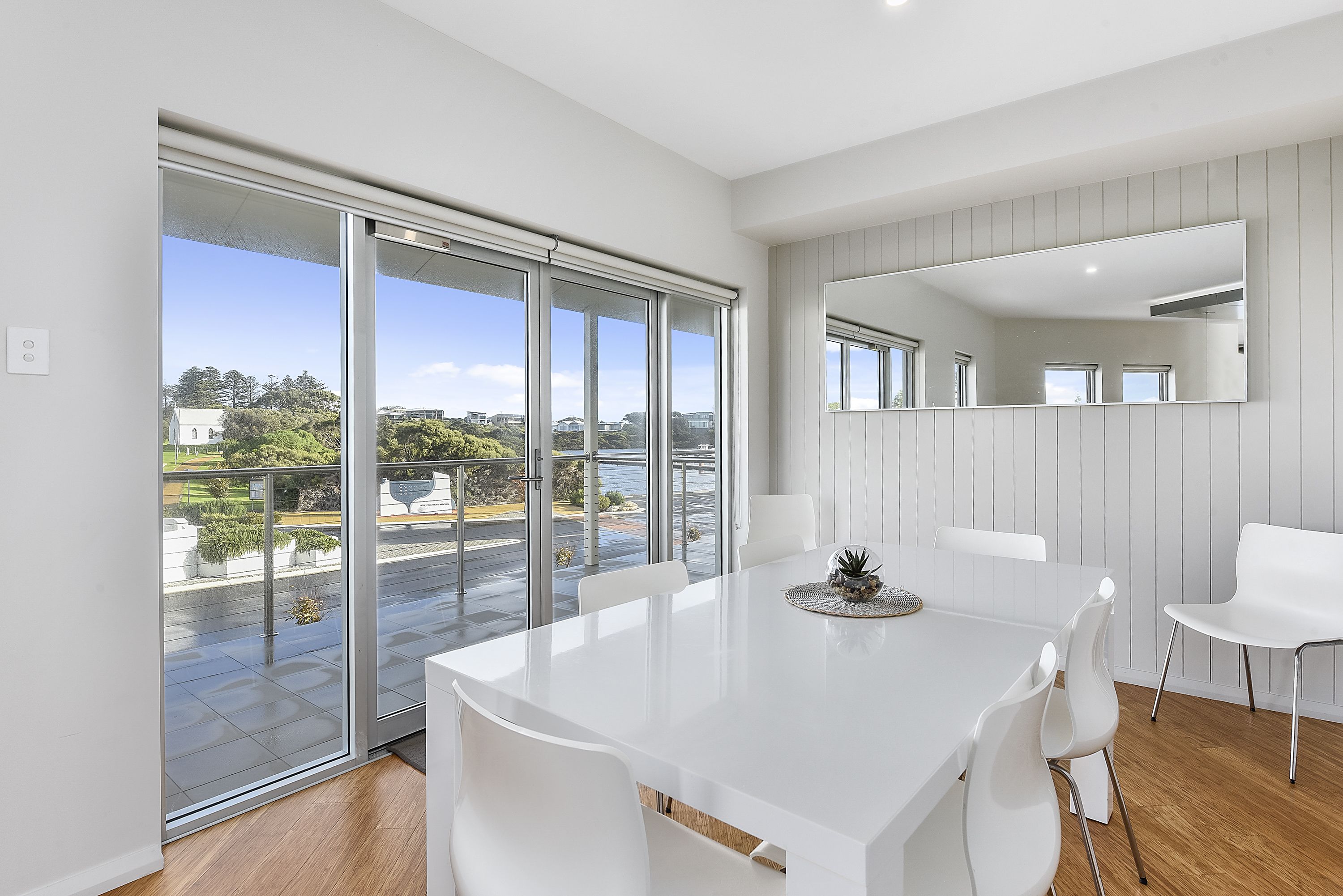 A modern dining area with a white table and chairs, large sliding glass doors leading to a balcony with a city view.