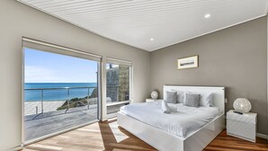 A bedroom with a large bed, a nightstand, and a view of the ocean through a sliding door. - Aloha Waterfront Properties (Robe)