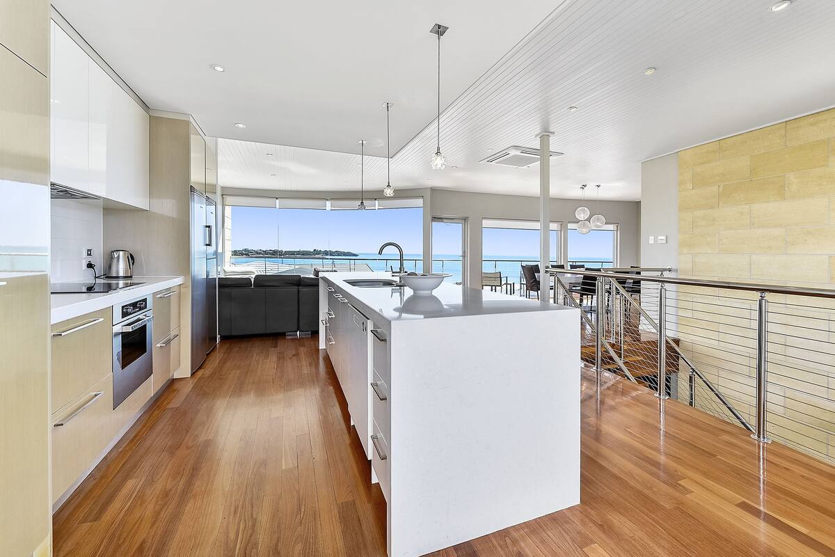A modern kitchen with a large island, wooden flooring, and a view of the sea.