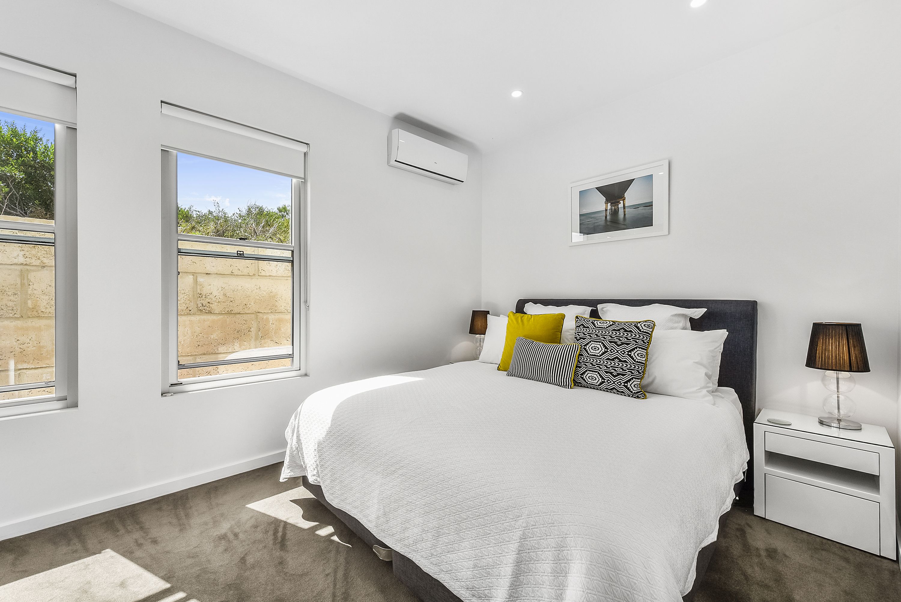 Un dormitorio moderno con una cama grande, una mesita de noche, aire acondicionado instalado en la pared y una ventana con vistas a zonas verdes.