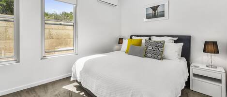 Un dormitorio moderno con una cama grande, una mesita de noche, aire acondicionado instalado en la pared y una ventana con vistas a zonas verdes.