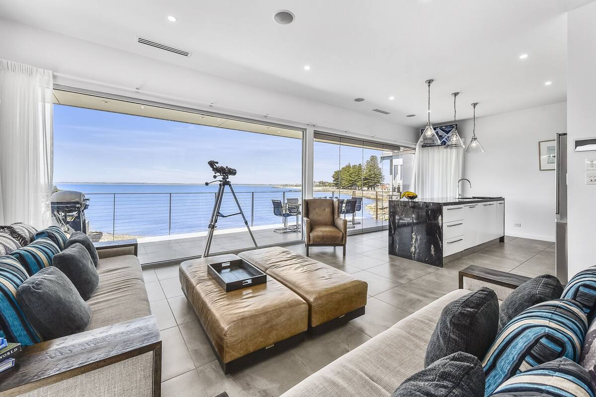 A modern living room with a large window overlooking the sea, a sofa, ottoman, and a tripod-mounted camera.