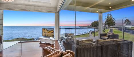 A modern living room with a glass wall offering a view of the sea and a balcony with outdoor furniture.