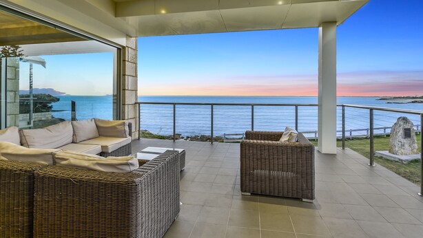 A modern outdoor seating area with wicker furniture and a glass wall overlooking the sea. - Aloha Waterfront Properties (Robe)