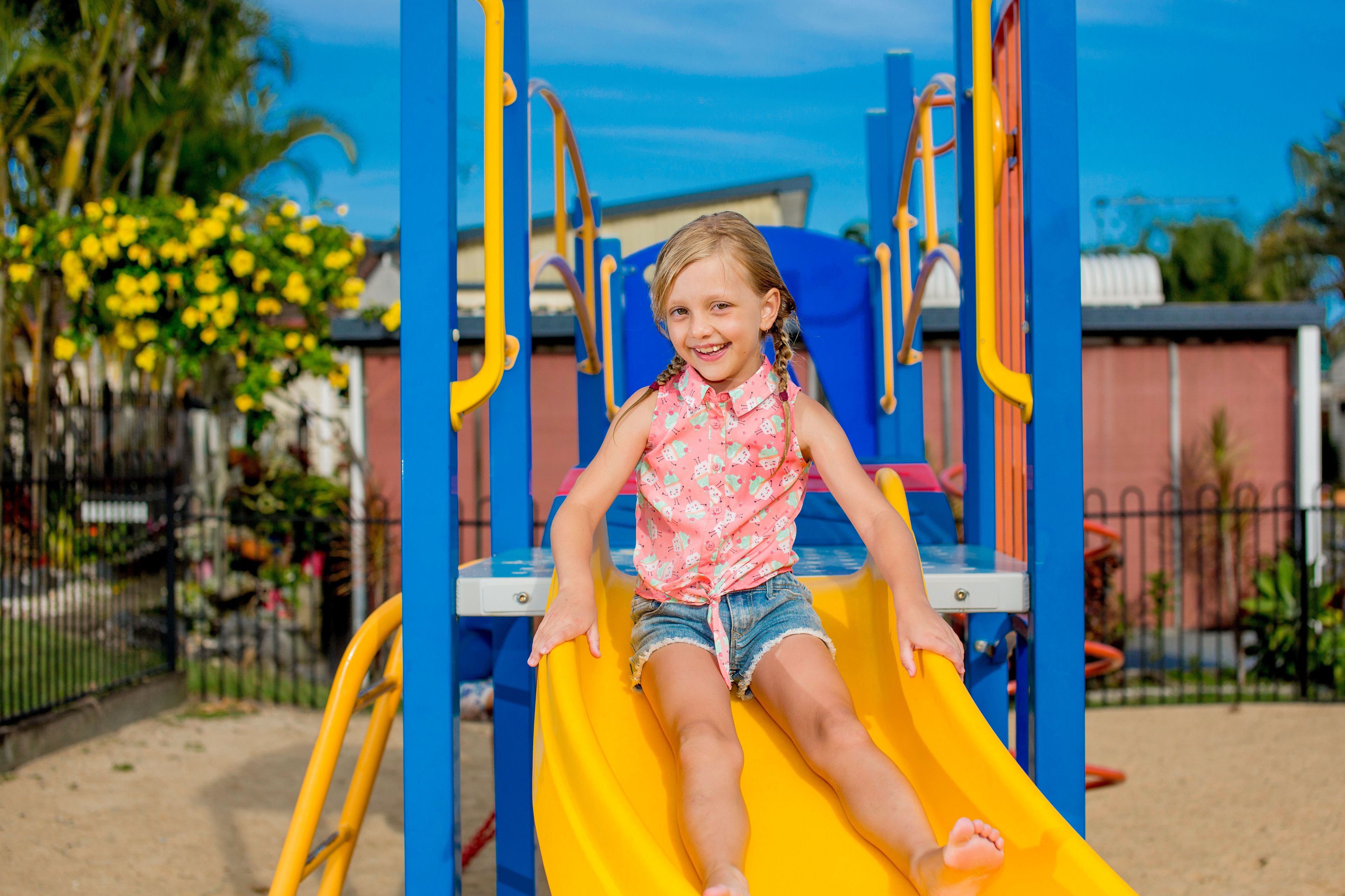 Un niño en un tobogán amarillo en un parque infantil con juegos azules y amarillos.
