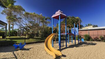 A playground with a slide and climbing structures, surrounded by a fence and palm trees.