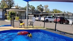 A swimming pool with a blue shade sail overhead, surrounded by a fence and a few chairs. - Silver Oaks Motel (Gilgandra)