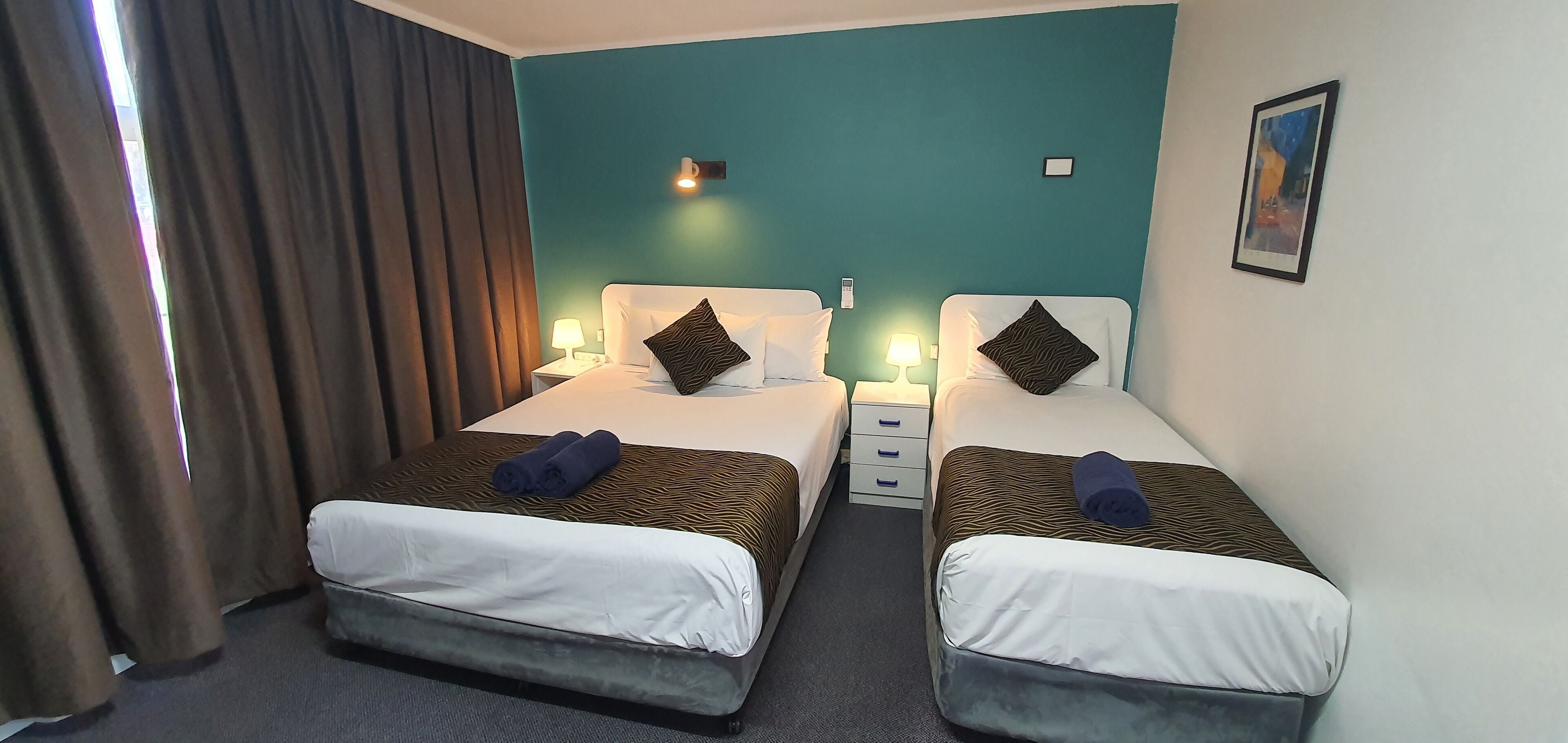 A hotel room with two beds, each with a pillow and a rolled towel. There are bedside tables with lamps and a wall-mounted light.