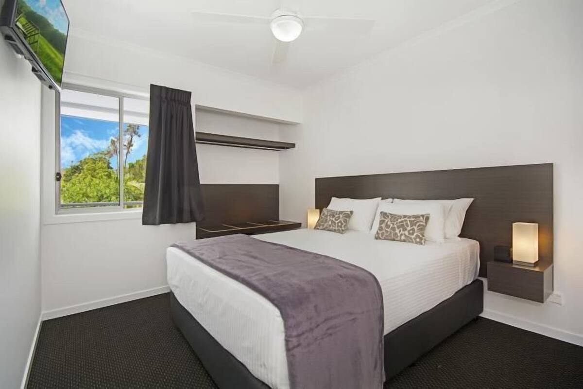 A hotel room with a large bed, a flat-screen TV mounted on the wall, a ceiling fan, and a window with a view of greenery.